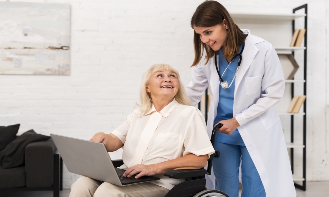 Na zdjęciu opiekunka medyczna ze starszą kobietą na wózku pomagająca jej z laptopem
